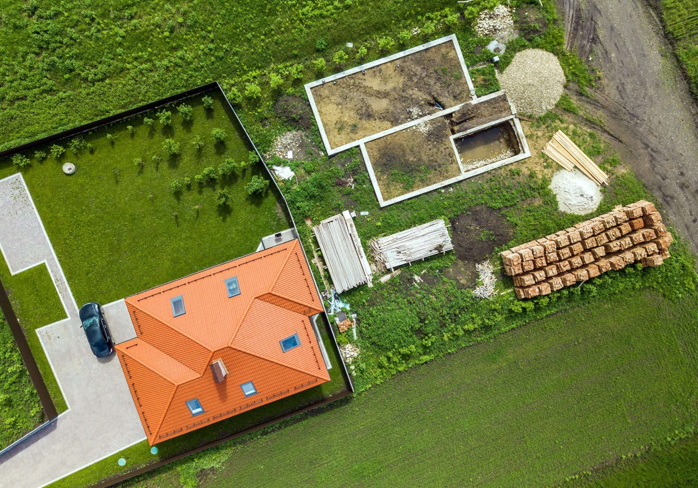 Aerial top view of a private house with attic windows on roof, paved yard with green grass lawn and building site with concrete foundation floor and stacks of yellow bricks for construction..
